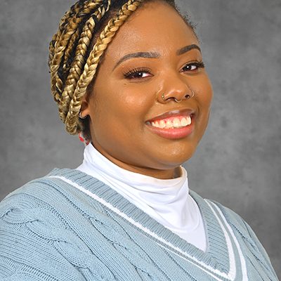 Jazz smiling at the camera. She has blonde braids and is wearing a blue sweater with a white turtleneck