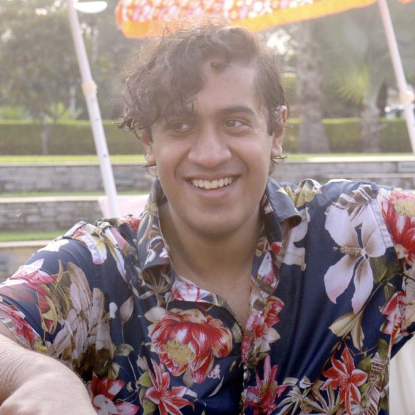 Adi is smiling at the camera. He has short dakr curly hair and is wearing a floral button up shirt.