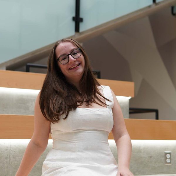 Anna is sitting on the stairs smiling. She has long brown hair and glasses and is wearing a white dress