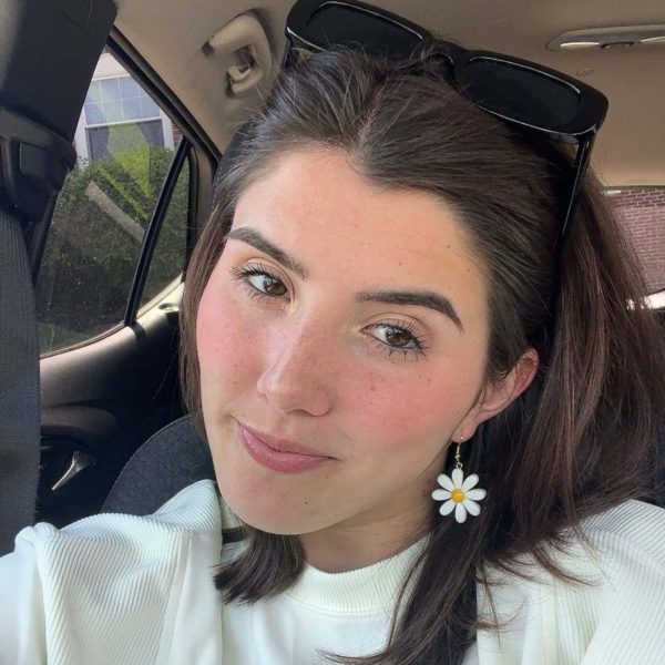 Chloe is sitting in the car. She has short dark brown hair and has daisy earrings and is wearing a white sweater