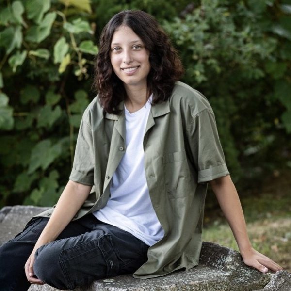 Emily is sitting on a rock outside. She has medium length curly brown hair and is wearing a white t shirt with an olive green button up.