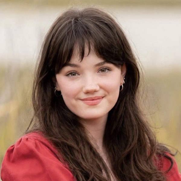 Eve is smiling at the camera. She has long dark hair and bangs and is wearing a red blouse sitting in a field.