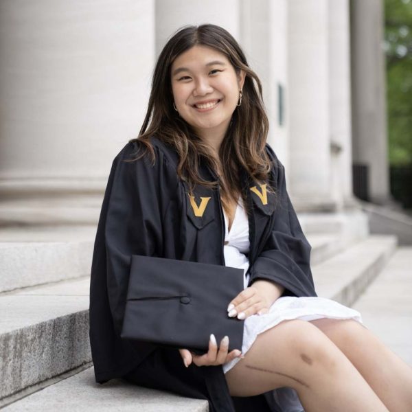 Fiona is taking graduation photos and is sitting on the stairs. She has long dark hair and is wearing a white dress with a black graduation robe and is holding her cap.