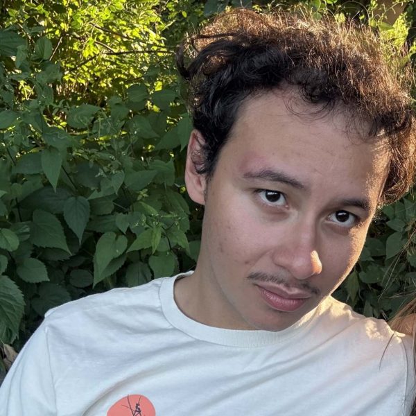 Gabriel is looking at the camera. He has dark curly hair and is wearing a white t shirt. He is sitting outside