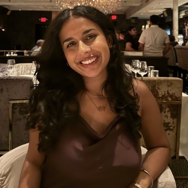 Gauri is smiling at the camera in a restaurant. She has long black wavy hair and is wearing a brown top