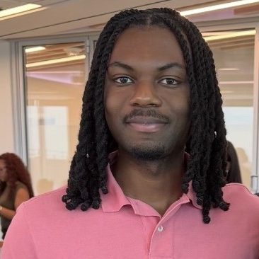 David is smiling at the camera. He has shoulder length dark hair in braids and is wearing a pink polo.