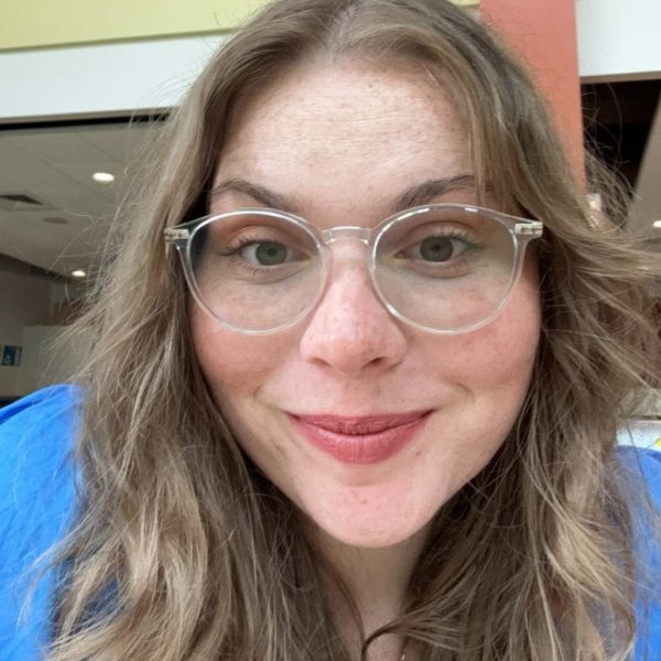 Jade smiles at the camera. She has long brown hair, glasses, and a blue top.