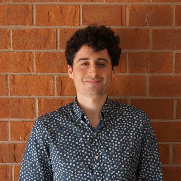Josh smiling at the camera against a brick wall. He is wear a blue button up shirt and has curly brown hair