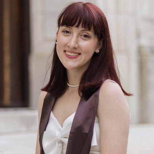 Lauren is posing for her graduation photo. She has medium length dark red hair and is wearing a white dress with a purple stoll.