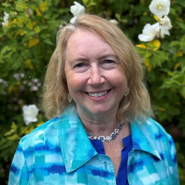 Marie smiling at camera in a garden with white flowers. She has shoulder length blonde hair and is wearing a teal jacket