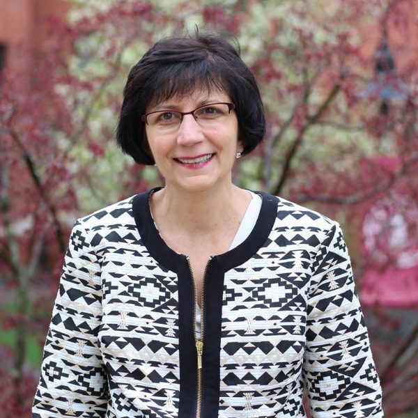Mary smiling at the camera. She has short black hair and glasses and is wearing a black and white patterned zip up sweater.