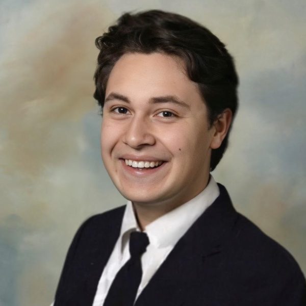 Matias is smiling at the camera. He has dark hair and is wearing a white button up with a tie and a jacket/.