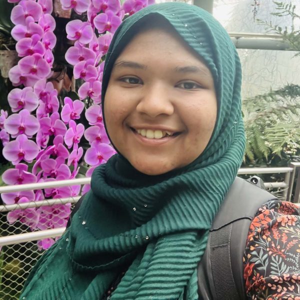 Roja is smiling at the camera. She has a green head scarf and is at a botanical garden with a big pink flower in the background