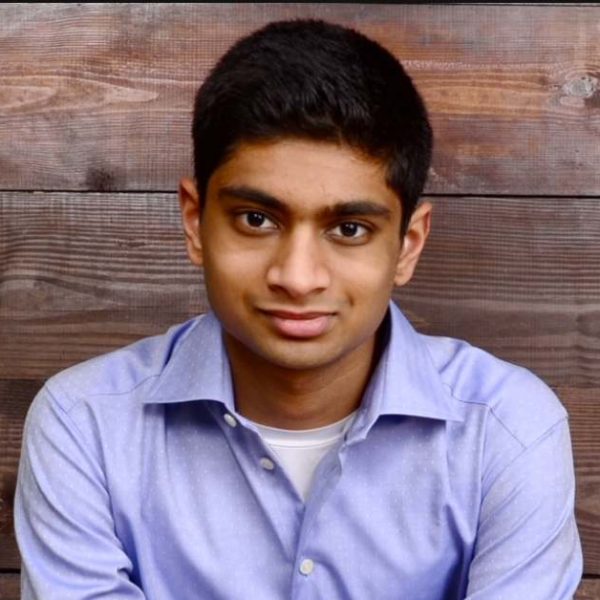 Neel is looking at the camera. He has dark hair and is wearing a light purple button up
