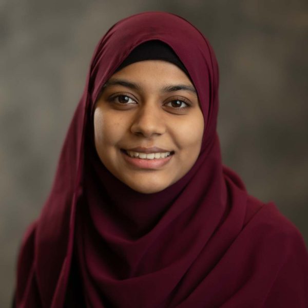 Zainab is smiling at the camera. She is wearing a dark red hijab with a gray background