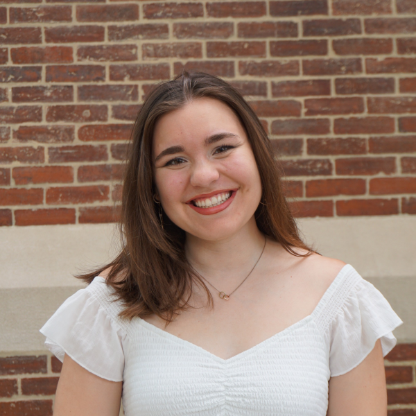 Courtney is smiling at the camera, wearing a white shirt, in front of brick background.