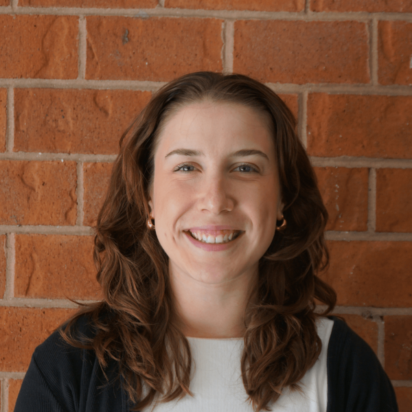 Photo of Sophia against a brick wall smiling at the camera. She has medium length brown wavy hair and is wearing a white top and black cardigan.
