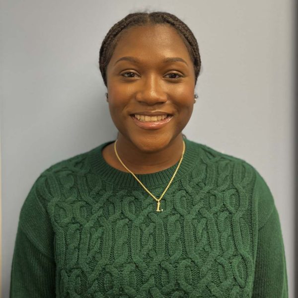 Lynsey looking at the camera smiling. She has black hair in a low bun, a green sweater, and a gray background.