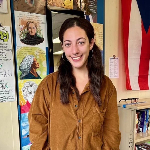 Makaila Cerrone looking at the camera smiling. She has long dark brown hair and is wearing a light brown suede button up and is standing in a classroom.